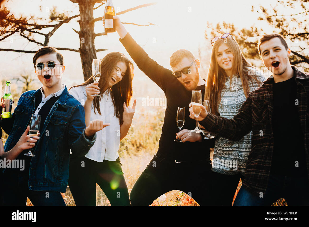 Group of attractive young people raising glasses and bottles with alcohol and yelling while standing in wonderful countryside. Friends screaming and r Stock Photo
