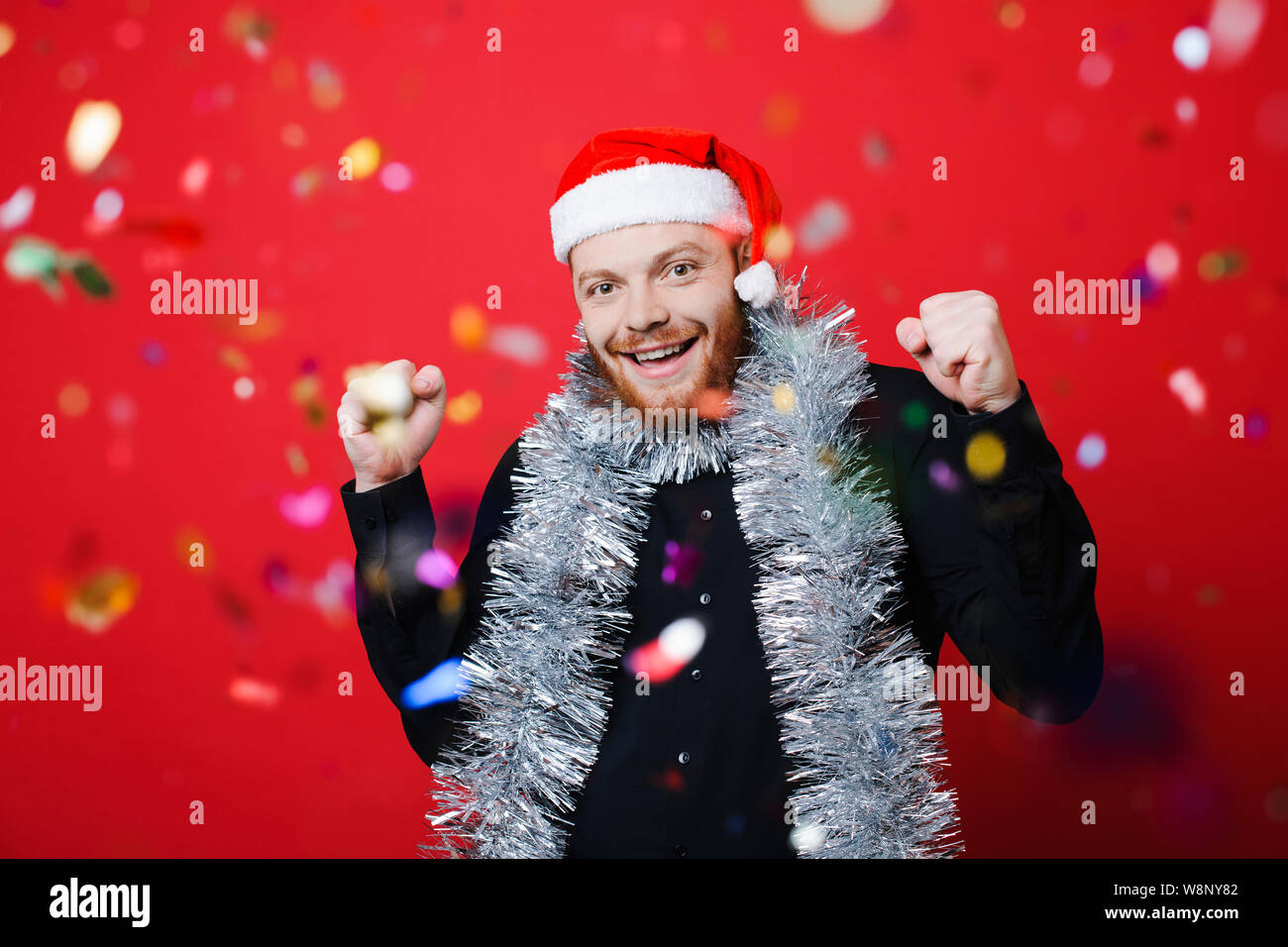Handsome man in Christmas hat gesturing with both hands and looking at camera while celebrating success and standing behind falling confetti on red ba Stock Photo