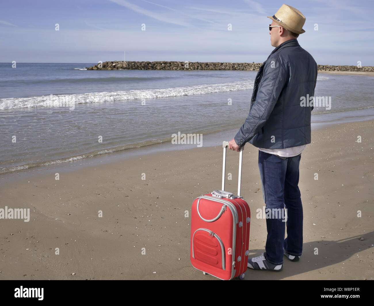 A man tourist in a hat and with a red suitcase stands on the seashore Stock Photo