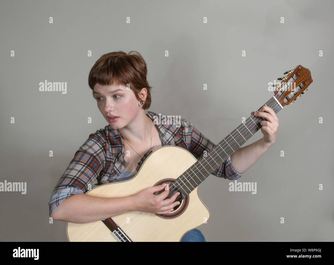A pretty young woman plays a classical guitar in a horizontal studio shot with a gray background. No brand ID is visible. Stock Photo