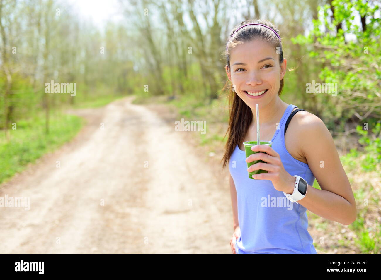 Healthy woman drinking green smoothie wearing smartwatch. Female runner resting drinking a spinach and vegetable smoothie using smart watch heart rate monitor during outdoor running workout in forest. Stock Photo