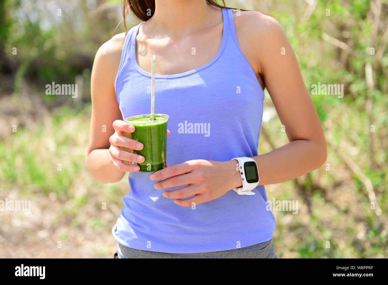 Green smoothie - woman runner wearing smartwatch. Healthy woman drinking vegetable smoothie wearing smart watch heart rate monitor during outdoor running workout in forest. Stock Photo