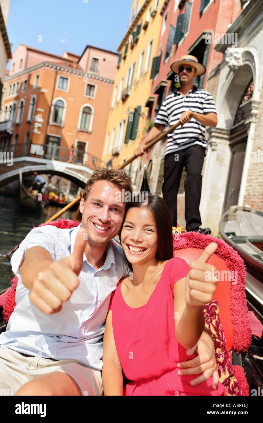 Travel. Happy couple traveling in Venice gondola giving thumbs up hand sign excited looking at camera. Romantic young beautiful couple on vacation holidays sailing in venetian canal in gondole. Italy Stock Photo