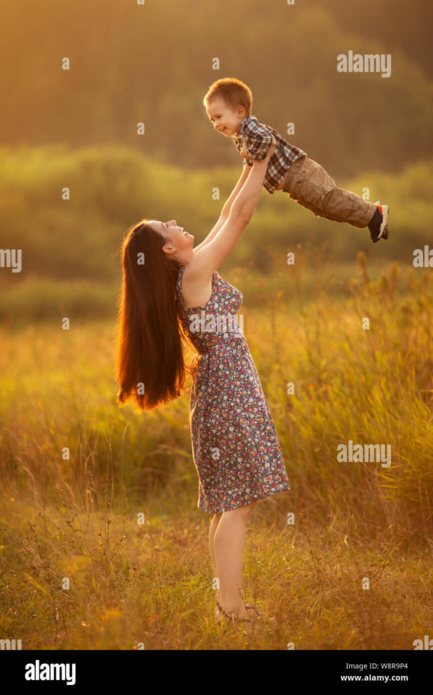 cheerful mother with toddler boy playing on the meadow on sunset. Beautiful woman lifting up her son outdoors. Happy family walking in summer nature Stock Photo