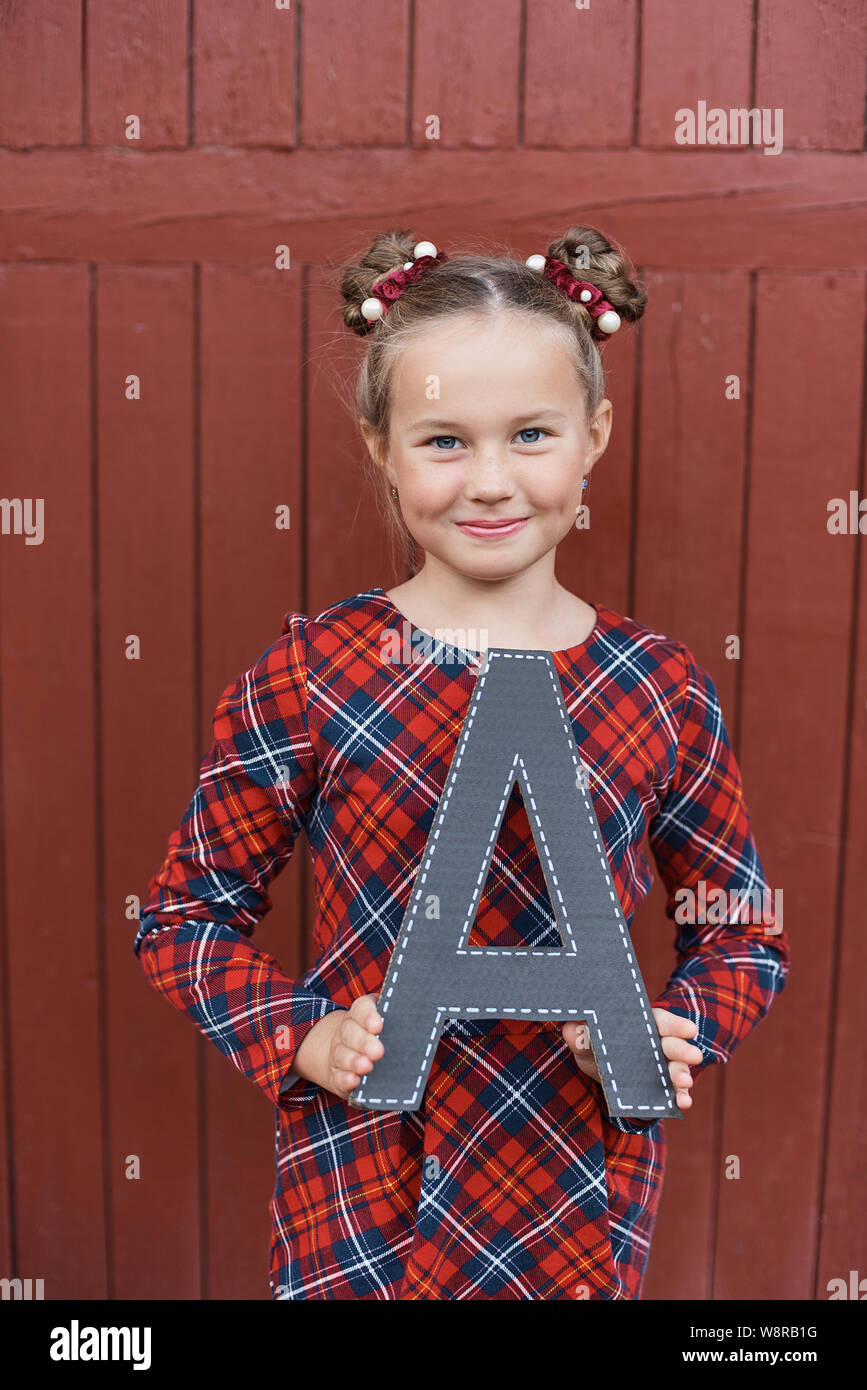 Portrait of a beautiful young Schoolgirl on the red wooden background witn big letter A. Farewell Bell. day of knowledge. beginning of the school year Stock Photo