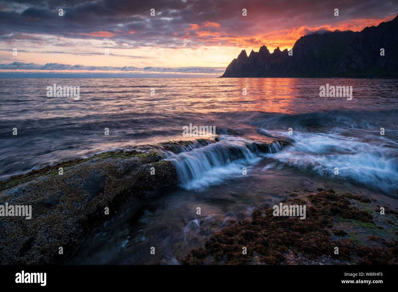 Waves crushing over mighty rocks with impressive oksen Mountains in background at a colorful midnight sunset, Tungeneset, Devils Jaw, Senja, Norw Stock Photo
