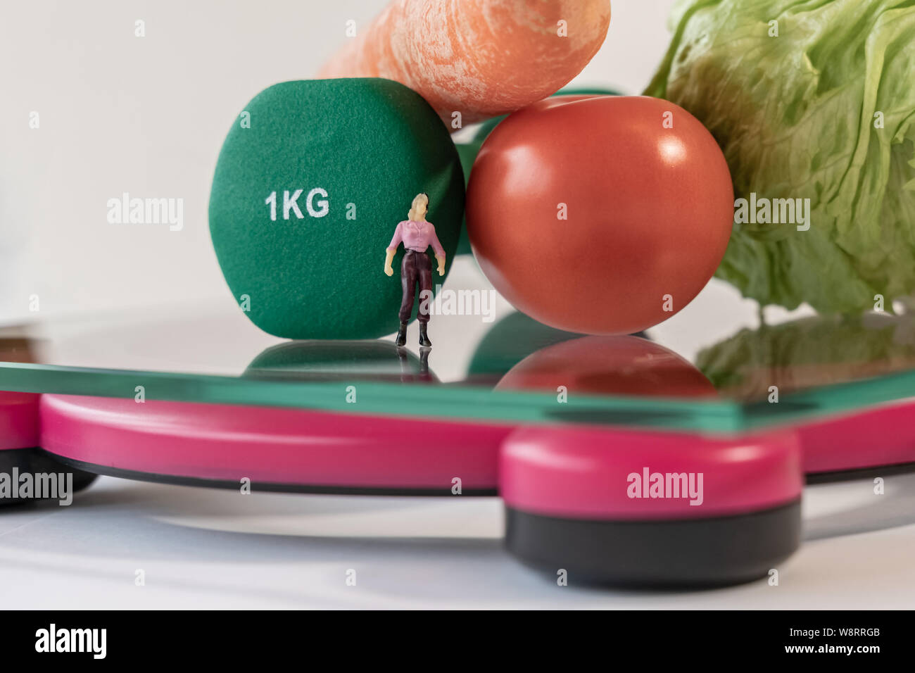Miniature woman figure standing on the digital electronic bathroom scale for weight of human body. Fresh vegetables and green dumbbells at shallow dep Stock Photo