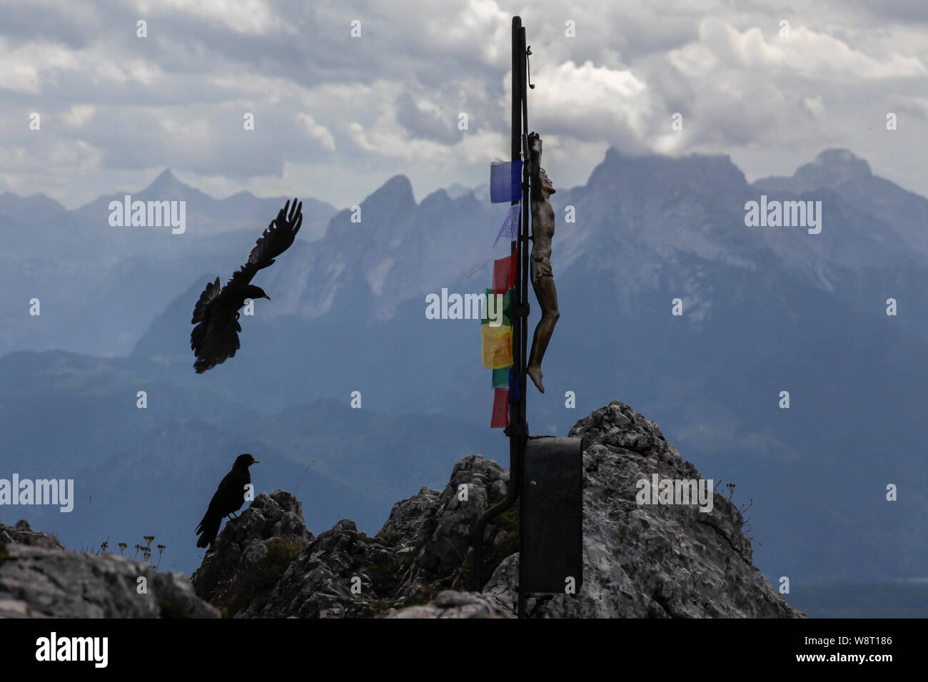 Two Yellow-billed Chough Pyrrhocorax graculus flying above a jesus cross on a mountain summit or ridge Stock Photo