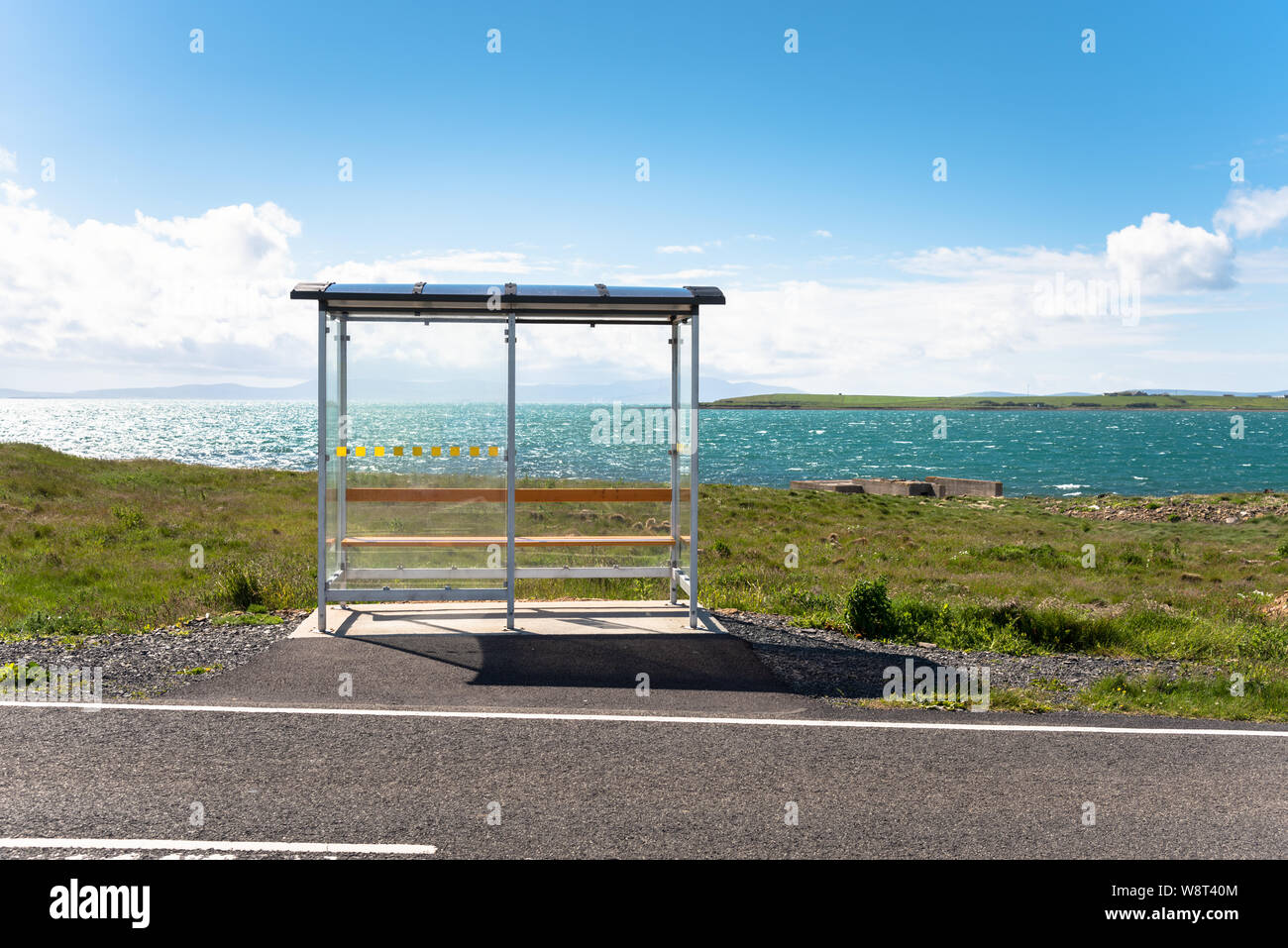 Empty bus stop with a glass shelter in a remote location along a coast road on a clear summer day. Scenic bay in background. Stock Photo