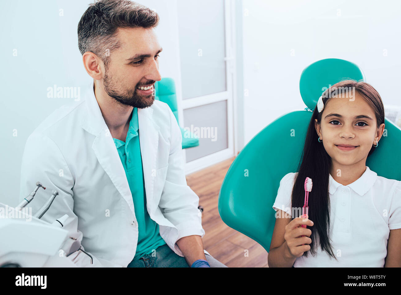Little mixed race girl in dental chair sitting near her dentist. She holding toothbrush and smiling. The Right Way to Brush Teeth Stock Photo