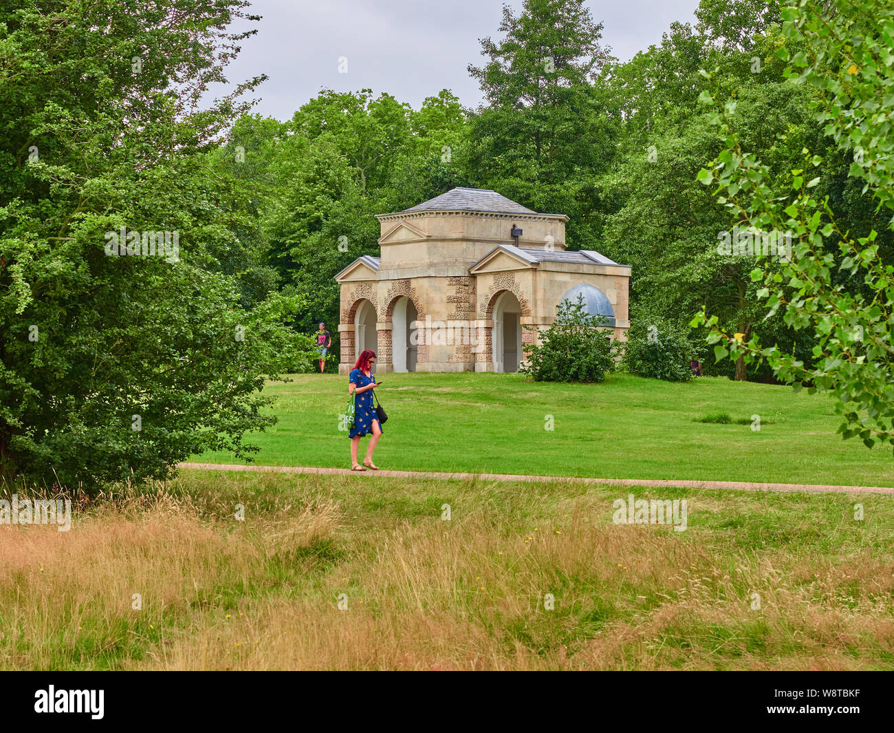 A red headed young woman in a blue dress walking in front of Queen Carolines Temple in Kensington Gardens whilst using her mobile phone Stock Photo