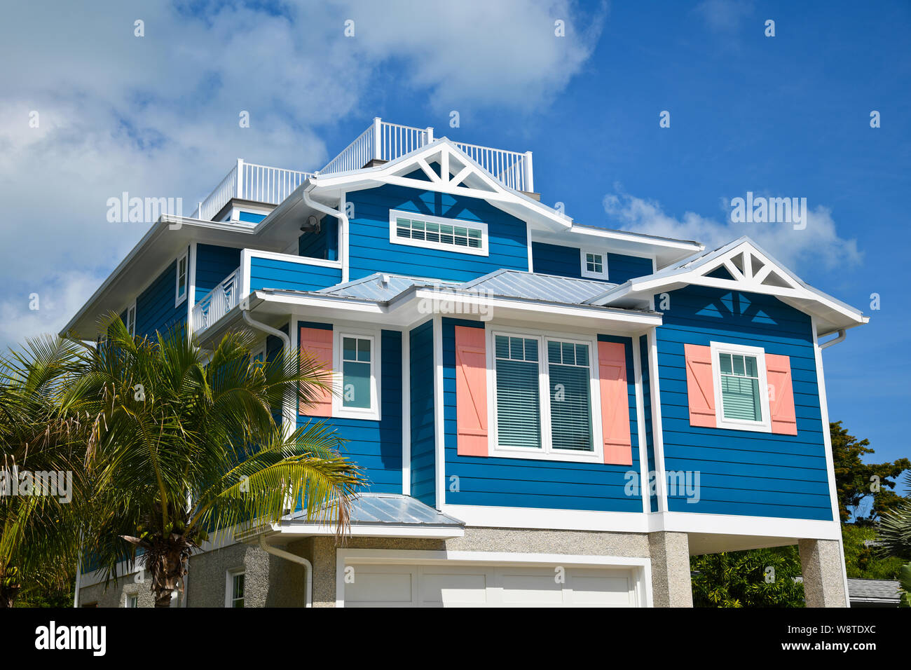 Large Beach House painted in dark blue with white trim and coral shutters. House has roof deck, garage and beautiful landscaping including Palm Trees Stock Photo