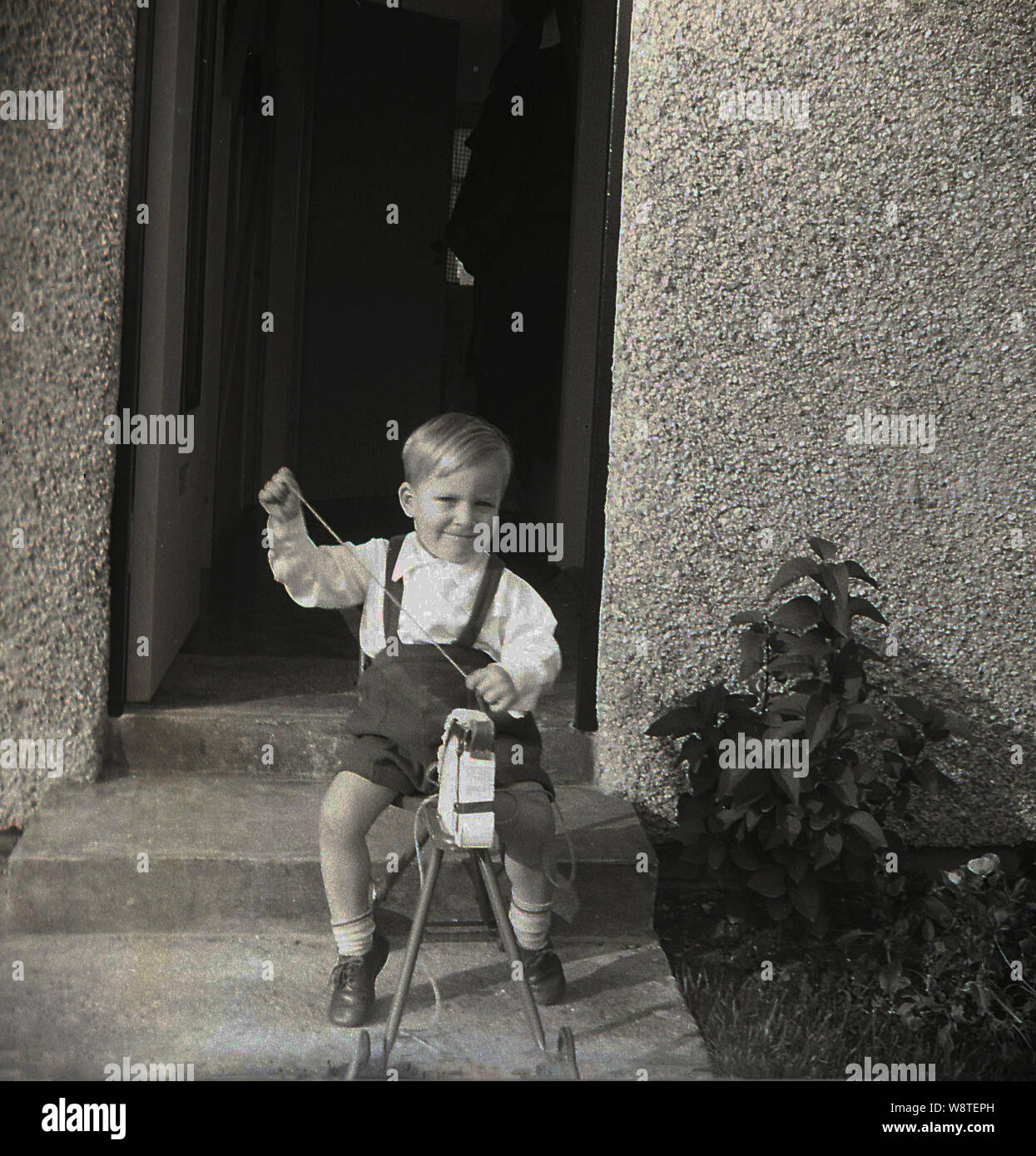 1950s, historical, a little boy outside a backdoor of pebble-dash house sitting on a ride-on toy horse with metal legs and wheels. Stock Photo