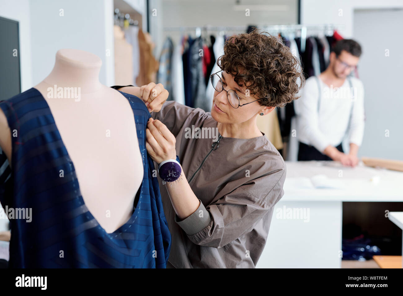Young serious seamstress stitching unfinished blue dress on mannequin Stock Photo