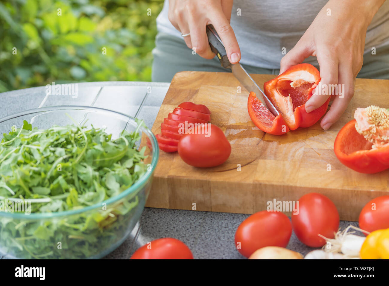 Closeup view of female hands slicing a red pepper on a wooden board. Various types of vegetables are lying on the table. Stock Photo