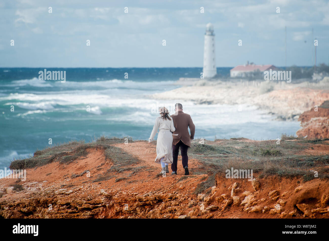 young pair man and woman in coat walking along stormy sea coast with beacon Stock Photo