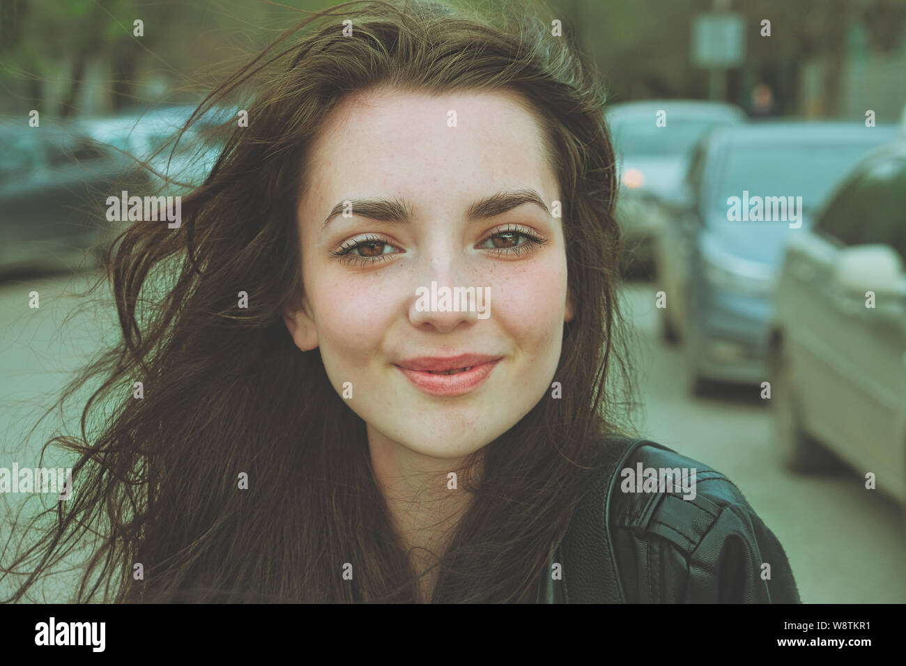 Happy girl posing outside in the street with parked cars on background looking at camera with windy hair Stock Photo