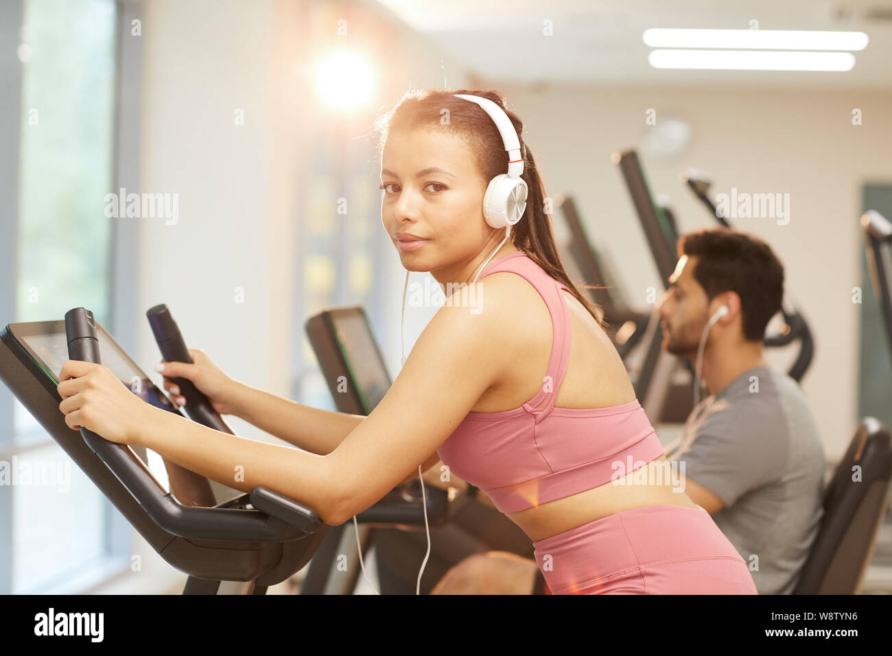 Waist up portrait of young woman wearing headphones looking at camera while exercising in gym, copy space Stock Photo