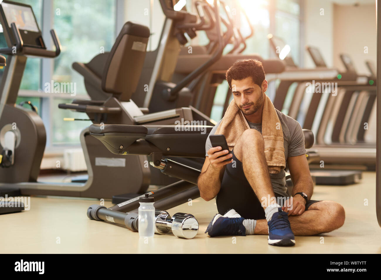 Full length portrait of handsome man resting sitting on floor in gym and using smartphone, copy space Stock Photo