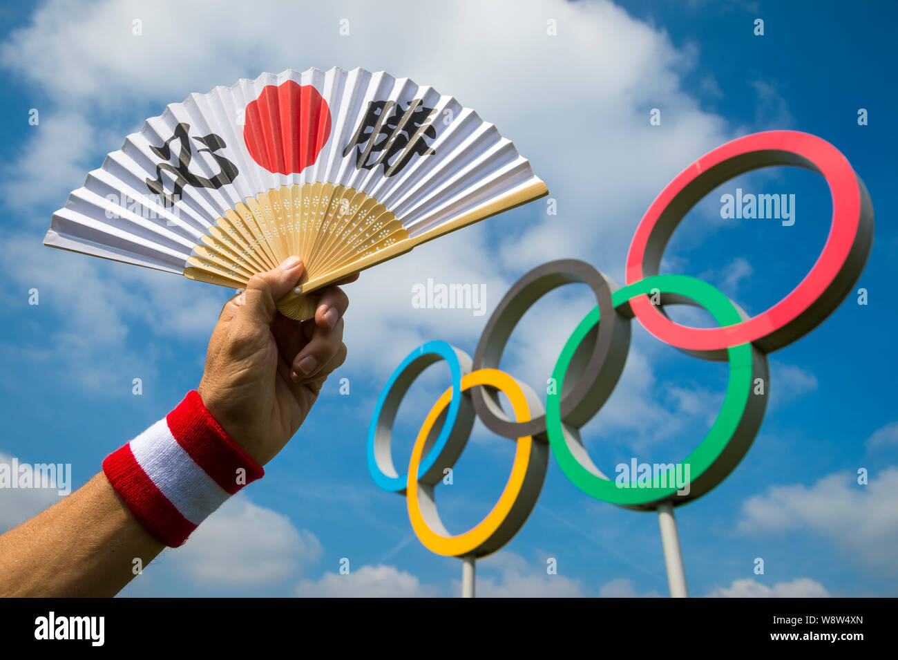 LONDON - APRIL 19, 2019: Hand holding a fan decorated with Japanese kanji characters spelling out hissho (certain victory) at a set of Olympic Rings. Stock Photo