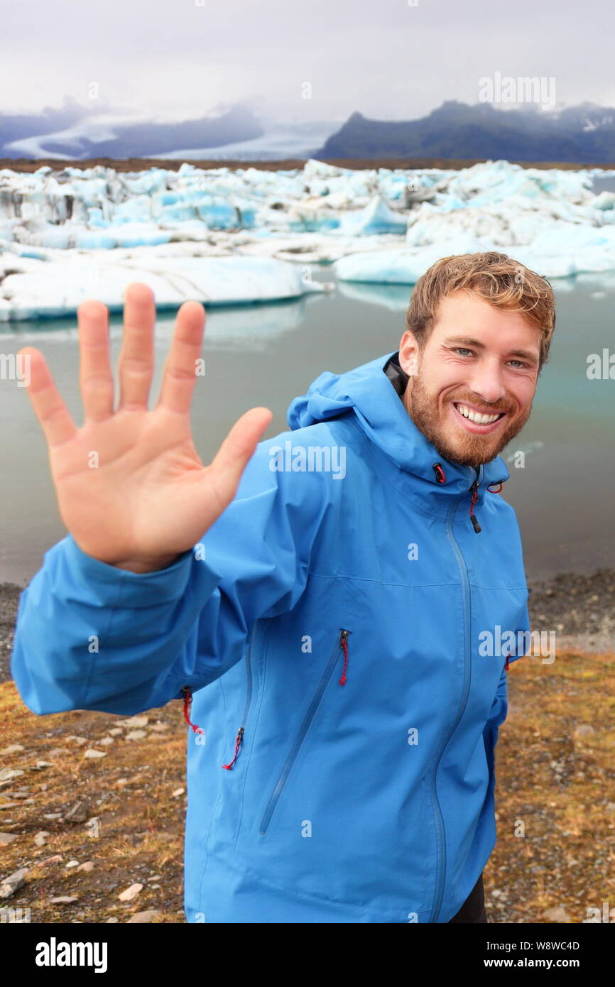 Tourist waving hand by Jokulsarlon on Iceland on travel. Portrait of happy man saying hello smiling looking at camera front of the glacial lake / glacier lagoon. Stock Photo