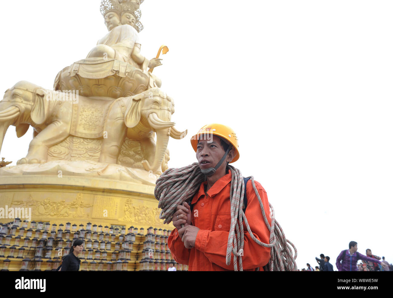 A Chinese cleaner carrying rope walks past the golden Buddha statue on the Golden Summit of Mount Emei (Emei Mountain) before climbing down a cliff to Stock Photo