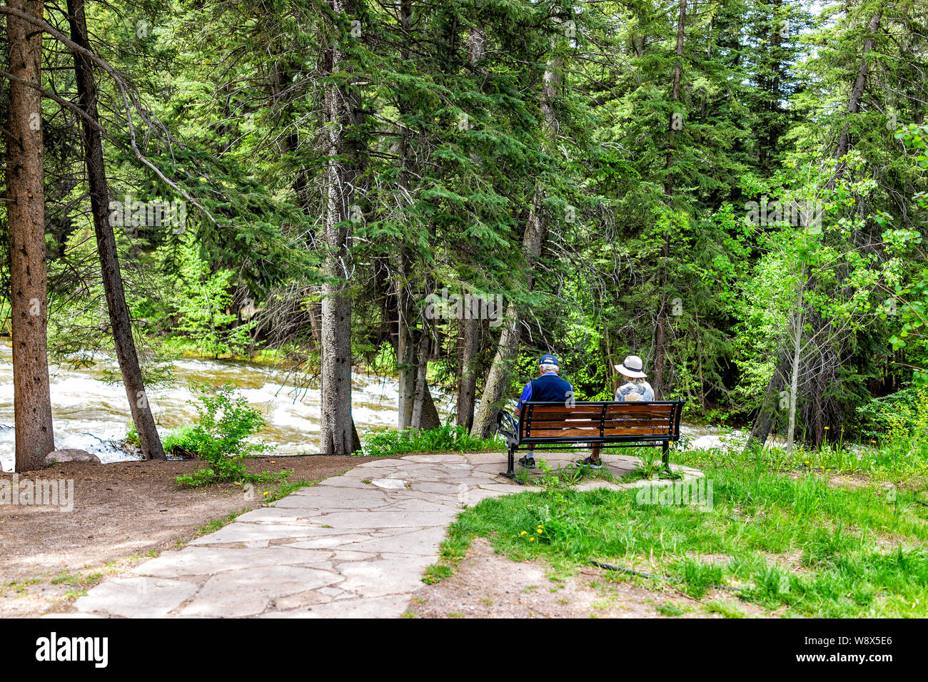 Vail, USA - June 29, 2019: Resort town in Colorado with senior couple sitting on bench by Gore creek river and pine trees Stock Photo