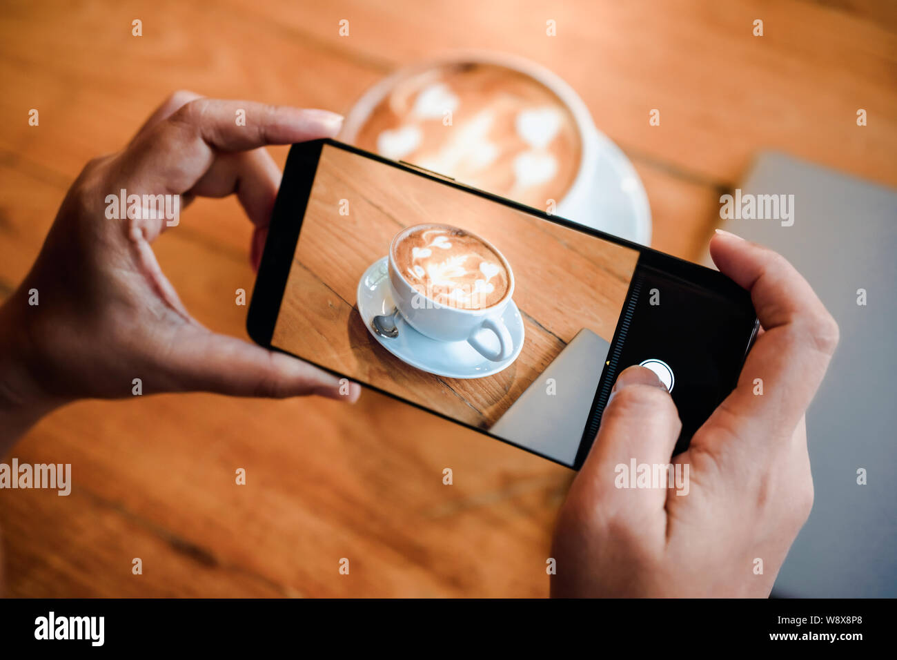 Hand taking a picture of hot latte art coffee cup with art foam on wooden table with her smart phone sitting at a coffee shop snap and share on social Stock Photo