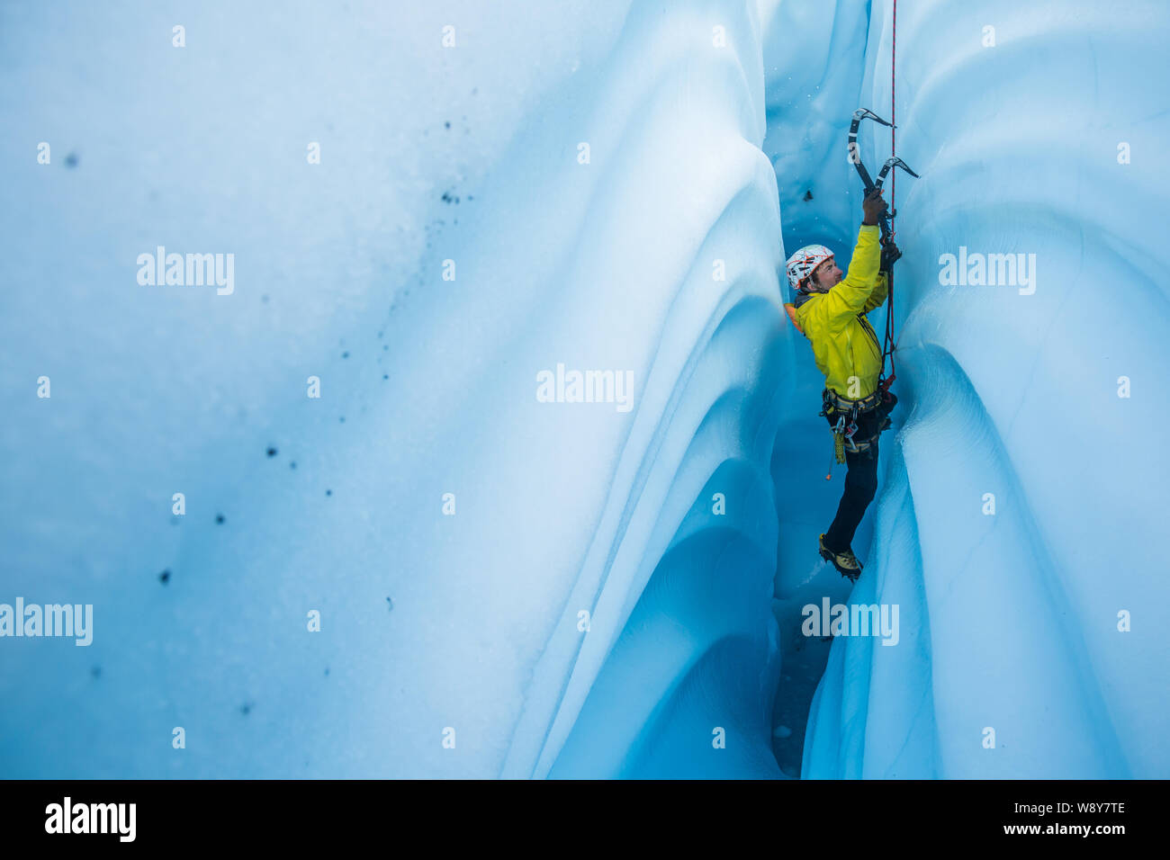 Man in yellow coat ice climbing a narrow and vertical canyon cut from solid glacier ice. Stock Photo