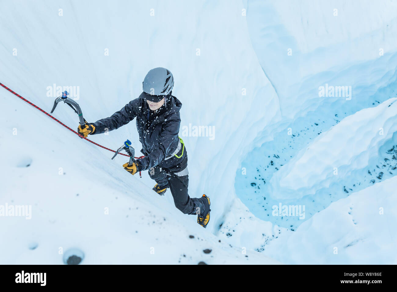 Woman smashes a tool into glacier ice as she climbs a steep pitch over a glacial river. The ice explodes into small pieces when the tool strikes it. Stock Photo