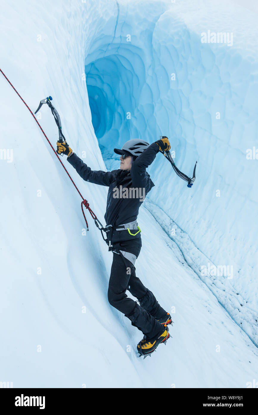 Ice climber in front of a massive ice cave in Alaska. She swings an ice tool behind her head as she climbs a vertical wall roped up from above. Stock Photo