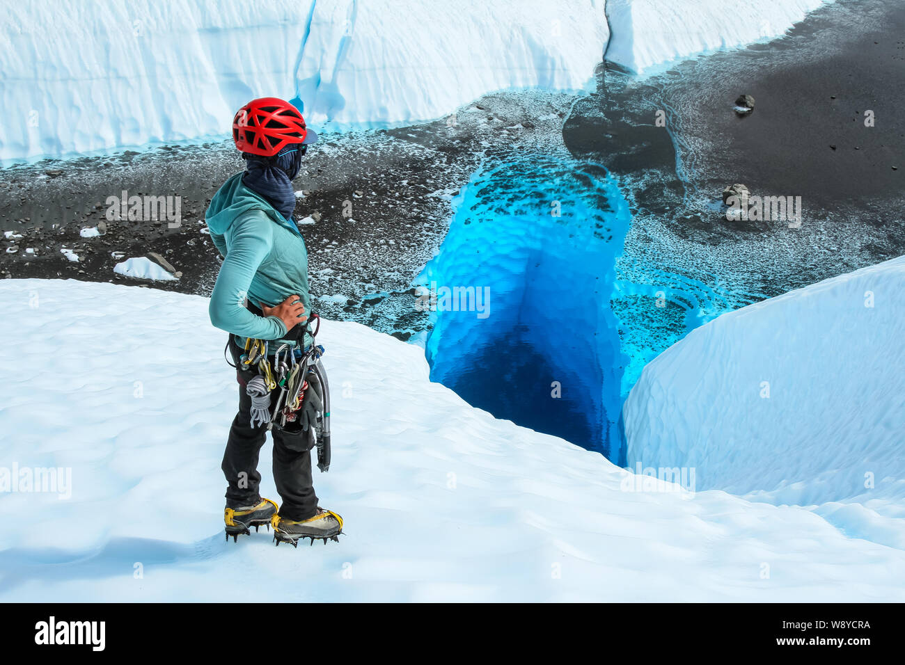 Woman in technical glacier gear and crampons stands over a deep blue hole looking into the clear water from above. Stock Photo
