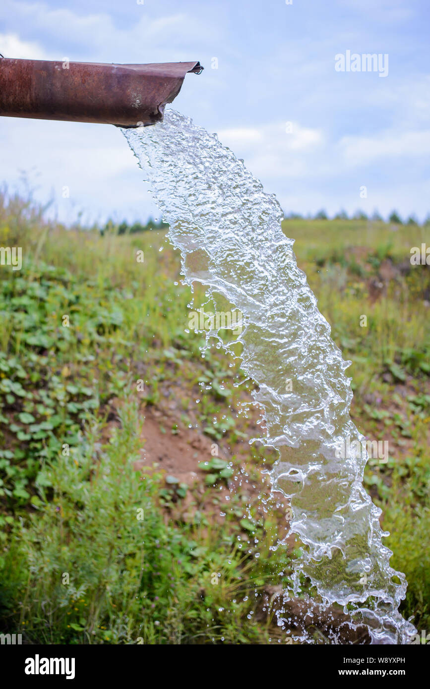 A strong stream of clean water pours out of a rusty old pipe in the countryside in the summer field Stock Photo