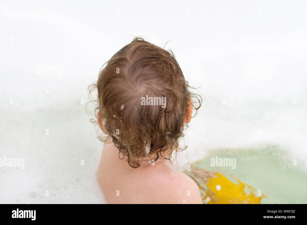 A small child is washing in the bath, baby girl boy in the water with soap suds, the view from the back only the head Stock Photo