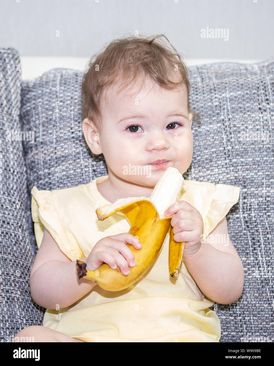 Little girl in yellow with a banana in her hand. Baby girl cleans ...