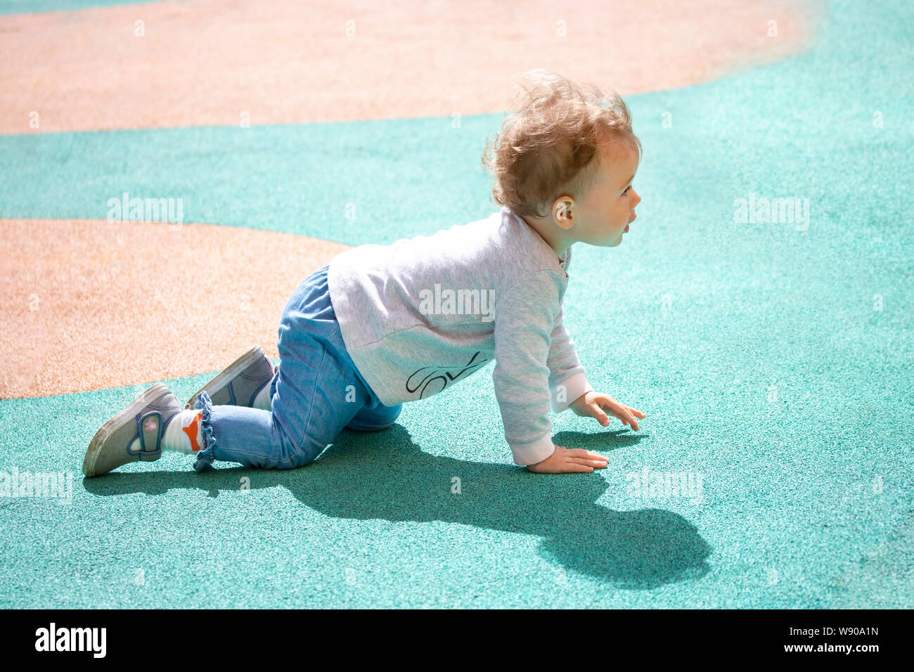 A 1 year old baby crawls around the playground for a walk. The kid is dressed in jeans, sweaters and denim boots. Stock Photo