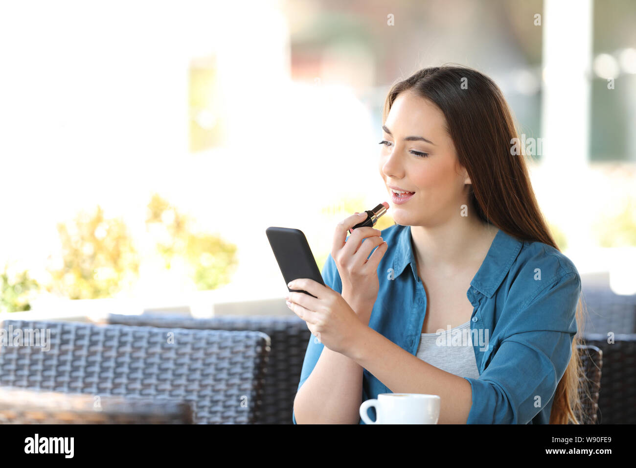 Woman painting lips using a phone as a mirror sitting in a coffee shop terrace Stock Photo