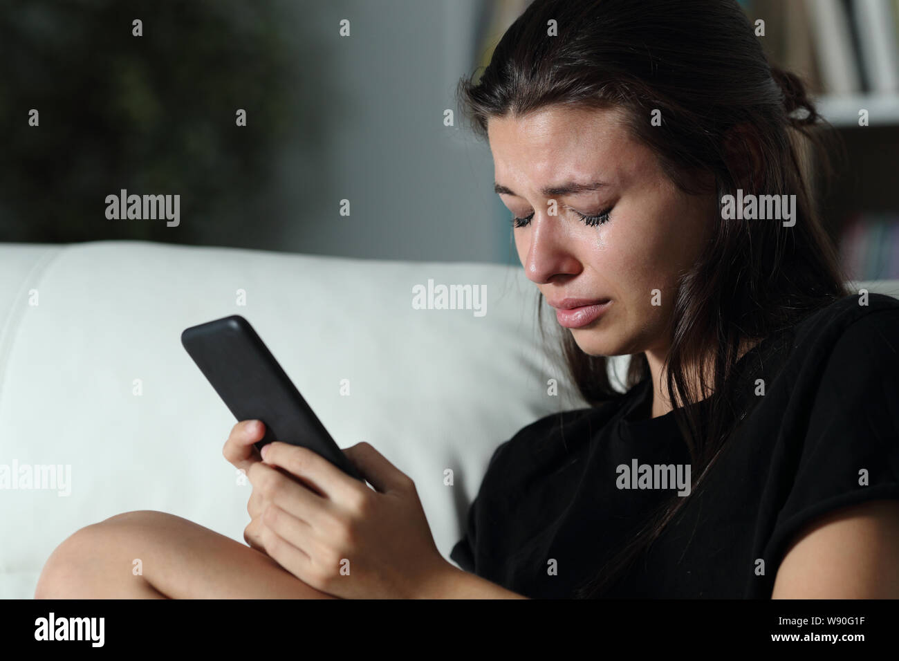 Sad teen crying after read smart phone message sitting on a couch in the dark at home Stock Photo
