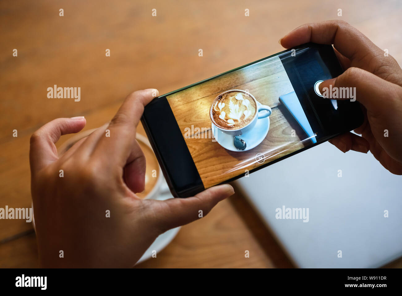 Hand taking a picture of hot latte art coffee cup with art foam on wooden table with her smart phone sitting at a coffee shop snap and share on social Stock Photo