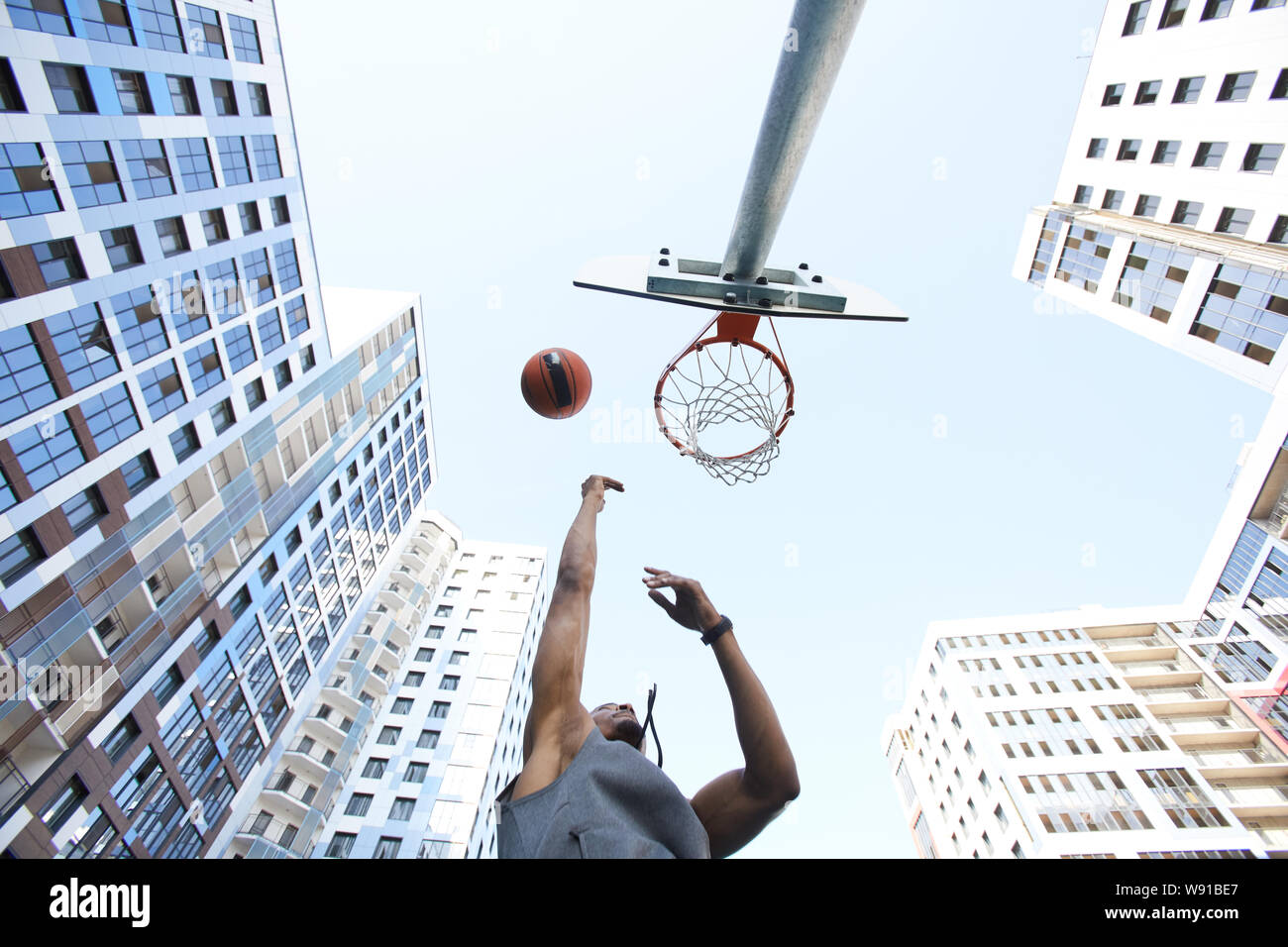 Low angle view at African basketball player shooting slam dunk against sky in urban background, copy space Stock Photo
