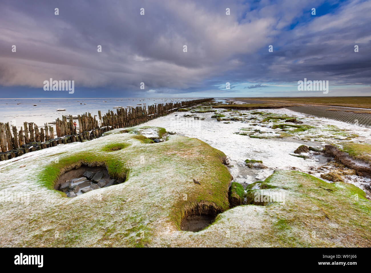 A winter landscape of the Waddensea in the Netherlands with dark clouds in the background and snow on the shore - Moddergat, The Netherlands Stock Photo