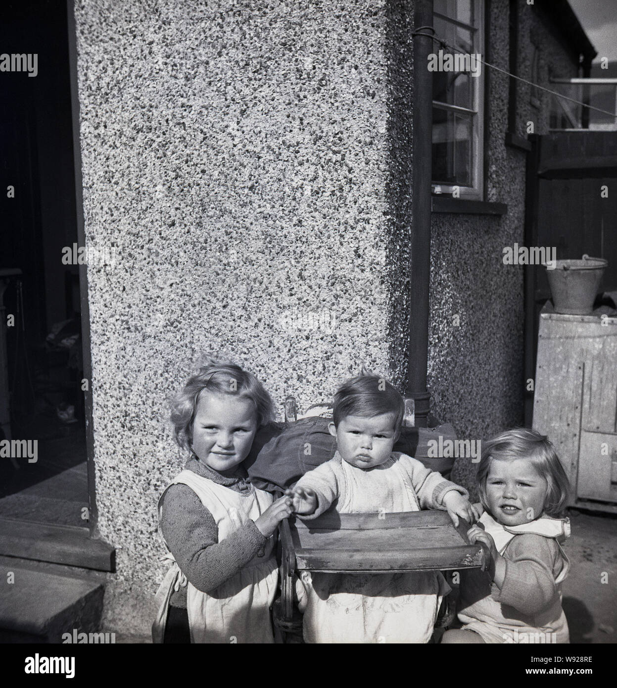 1950s, histroical, two young girls outside the backdoor of pebble-dash house, with their little infant sister sitting in a wooden high-chair, England, UK. Stock Photo