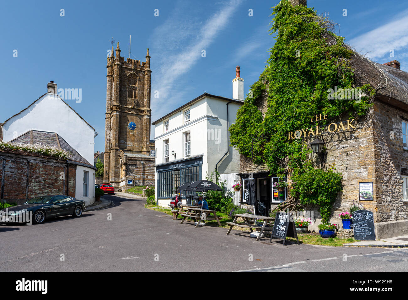 Cerne Abbas, England, UK - June 29, 2019: A man drinks outside The Royal Oak pub in the village of Cerne Abbas, with St Mary's Church behind. Stock Photo