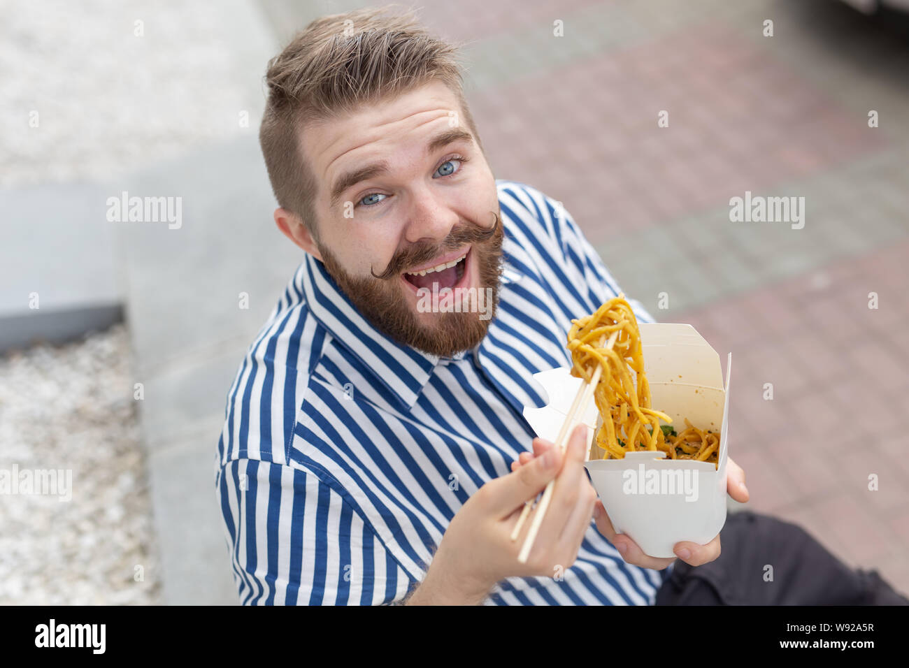 Close-up of an amusing young hipster guy eating chinese noodles with wooden  chopsticks sitting in a park outside on a warm summer day. The concept of  Stock Photo - Alamy