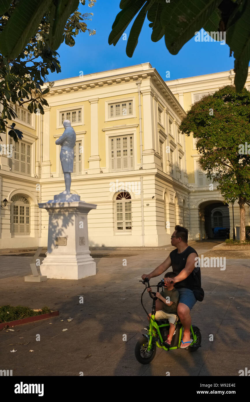 A cyclist and his son ride past the Raffles Statue, Raffles' Landing Site by the S'pore River, Singapore, the Museum of Asian Civilizations in the b/g Stock Photo
