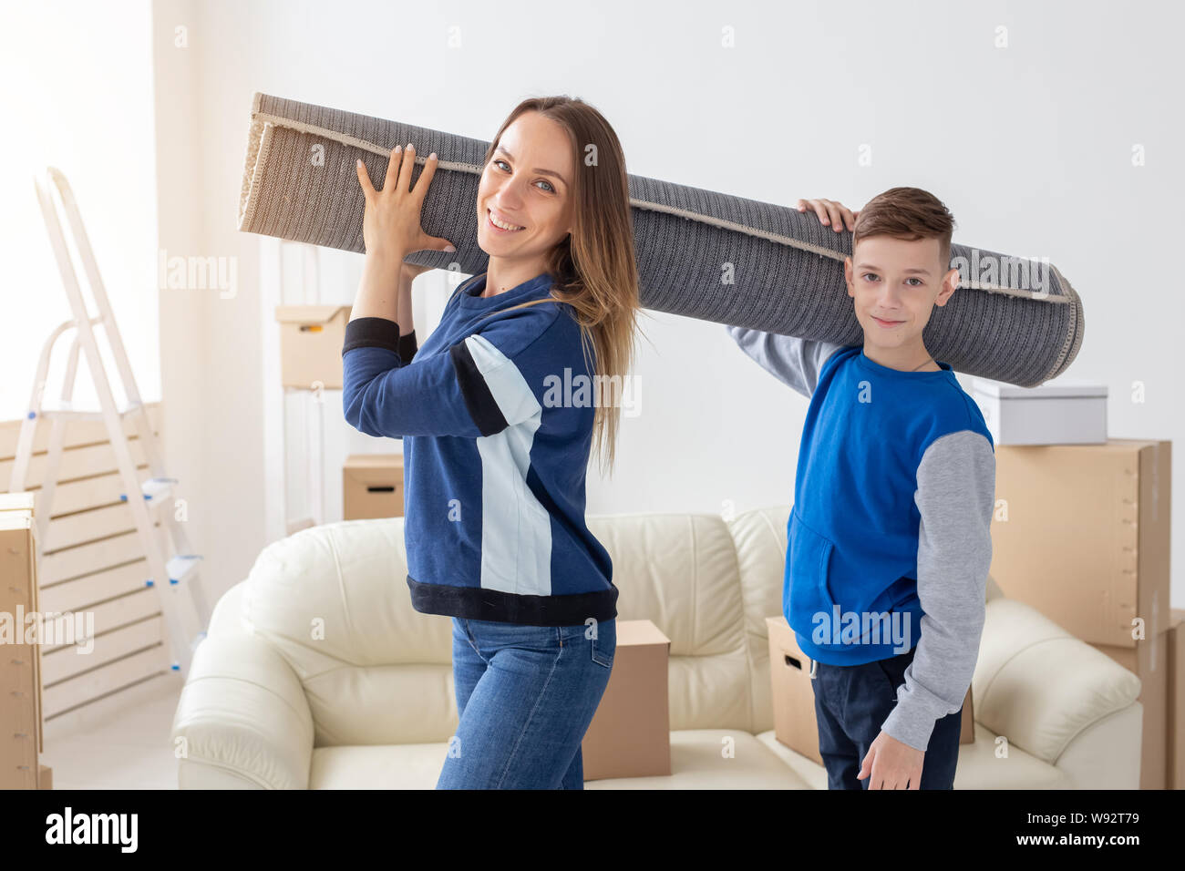 Smiling caucasian mother and a charming son are holding a folded carpet on their shoulders in a new living room intending to spread it in a new Stock Photo