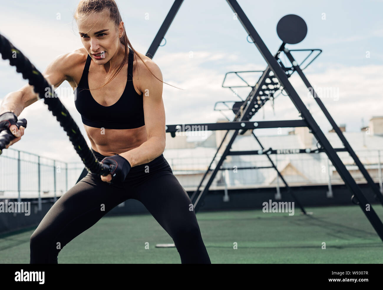 Woman using battle ropes during strength training on rooftop. Young female working out outdoors. Stock Photo