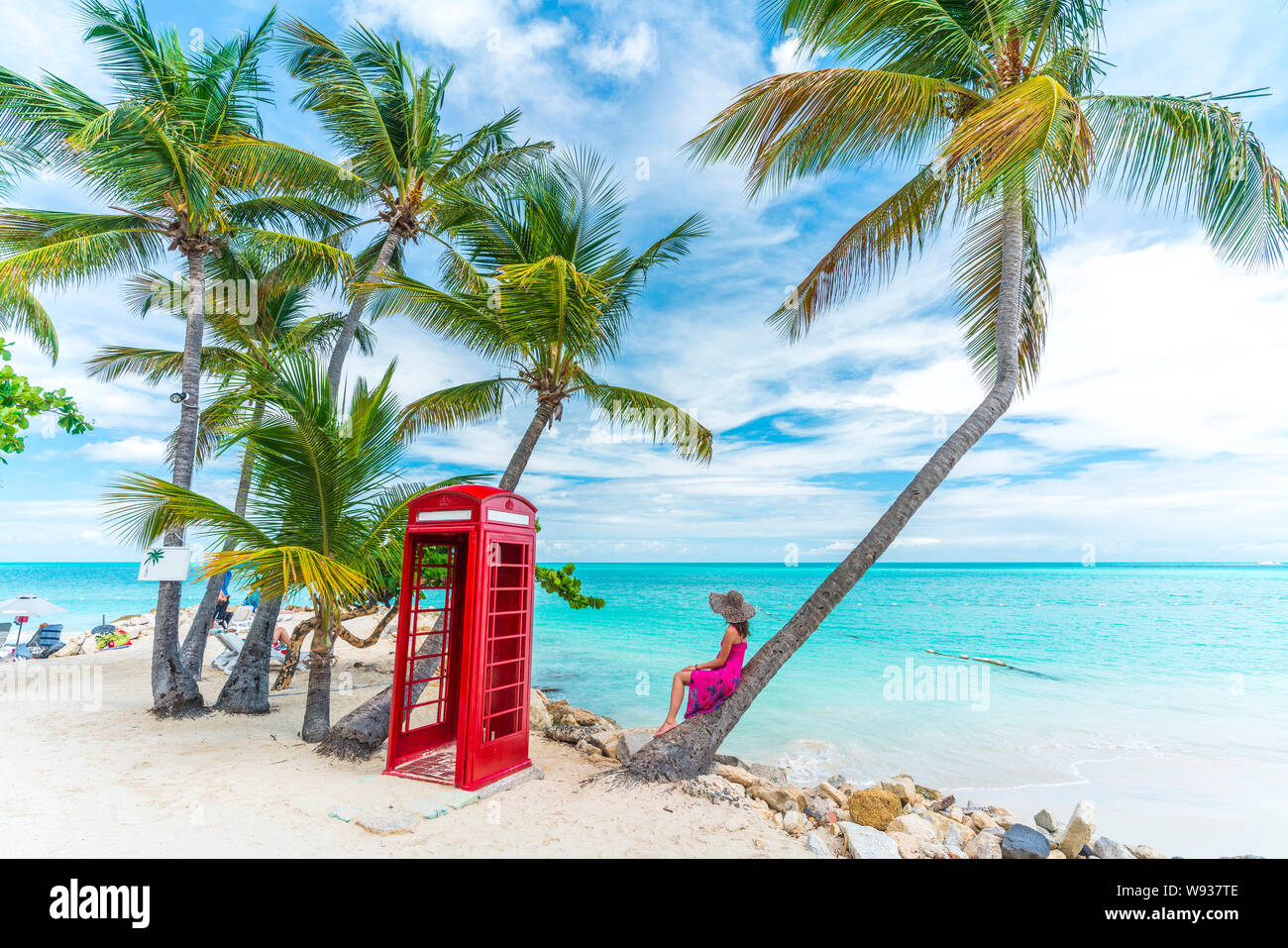 Woman sitting on palm tree beside the iconic phone box seafront, Siboney Beach, Dickenson Bay, Antigua, Caribbean Stock Photo