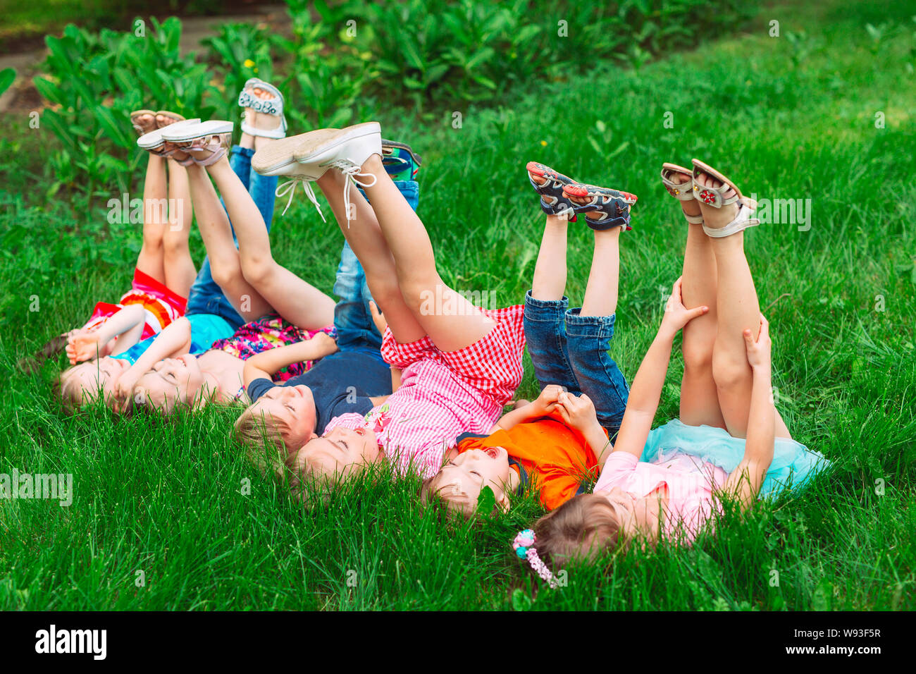 Children lying on green grass in park on a summer day with their legs lifted up to the sky. Stock Photo