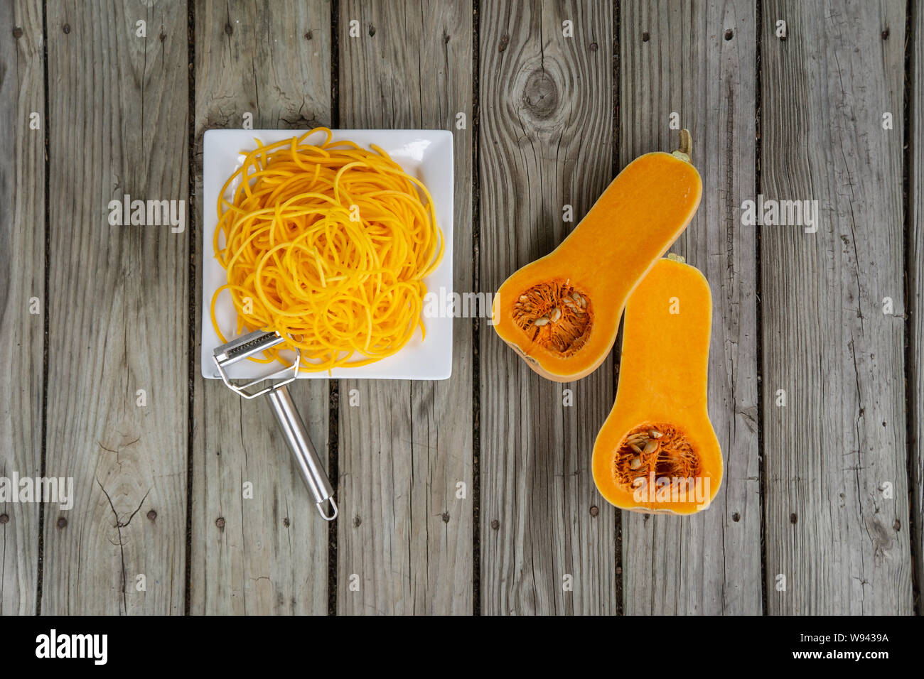 Overhead top view of freshly peeled butternut squash noodles and julienne peeler. Wooden background and white plate for fresh natural vegetable noodle Stock Photo